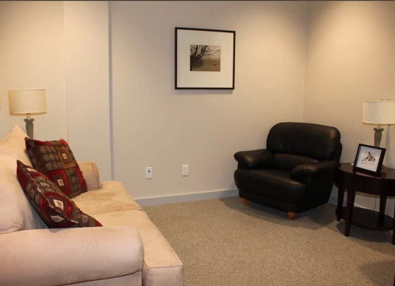 A modern living room with beige walls, cream-colored sofa with burgundy pillows, black armchair, side tables with lamps, framed artwork, and neutral carpeting