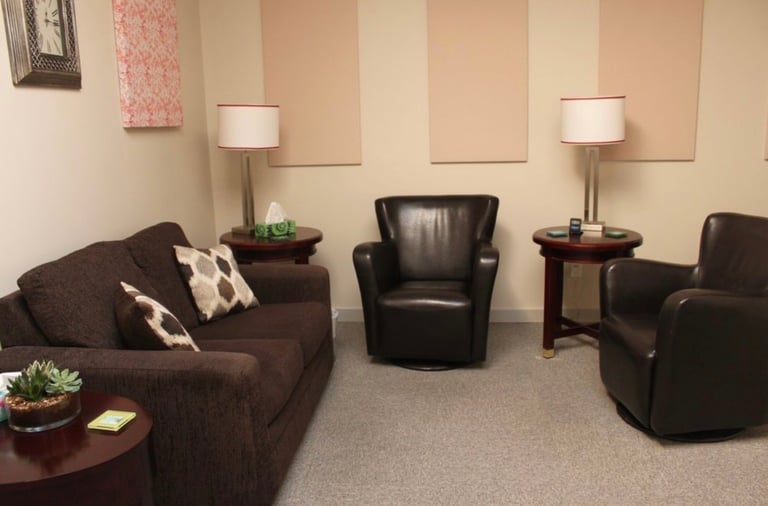 Modern waiting or therapy room with brown leather recliners, dark sofa with pillows, side tables with lamps, and neutral striped wall panels